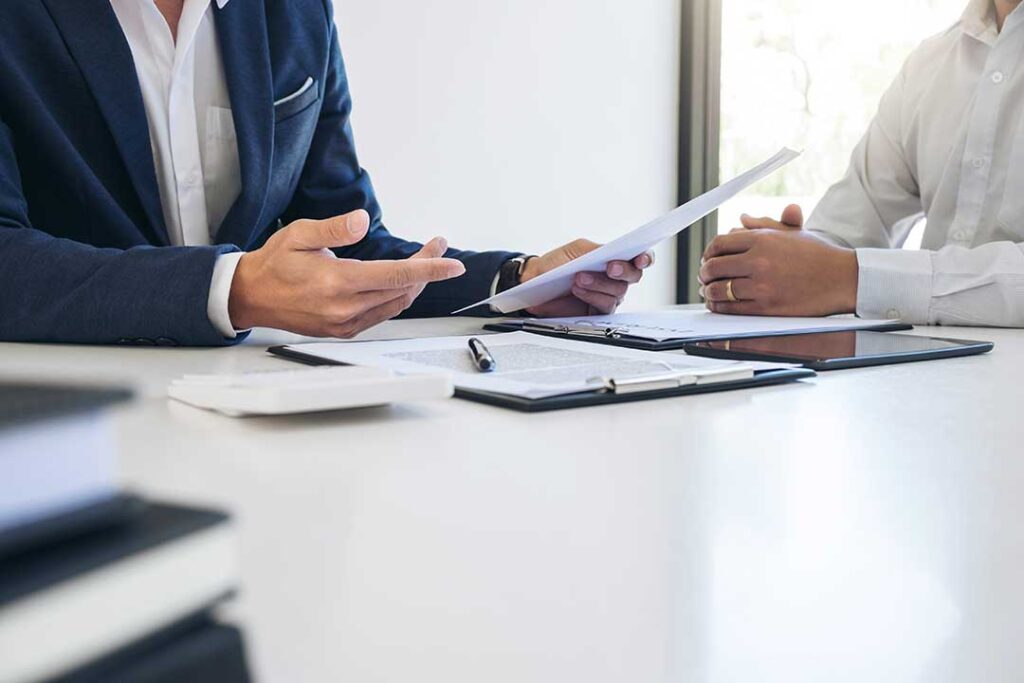 A person reviews paperwork and a calculator at a kitchen table with a car key and insurance policy documents.